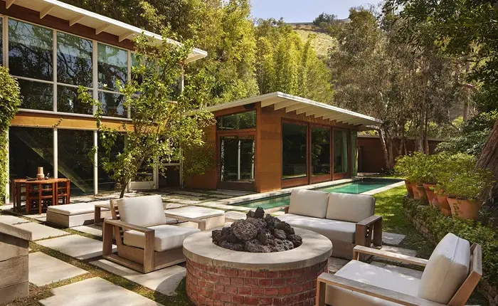 Modern backyard designed by architect Carl Maston, featuring a fire pit surrounded by cushioned patio chairs, a sleek lap pool, and a glass-walled house nestled amid trees and greenery