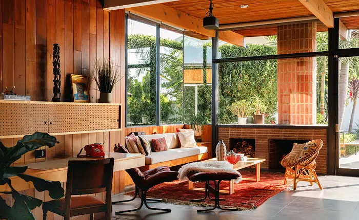 A cozy Mid Century Modern home living room in Jefferson Park features wood-paneled walls, large windows, a rattan chair, houseplants, and a brick fireplace—reflecting architect Thomas Downs’ signature style and an abundance of natural light.