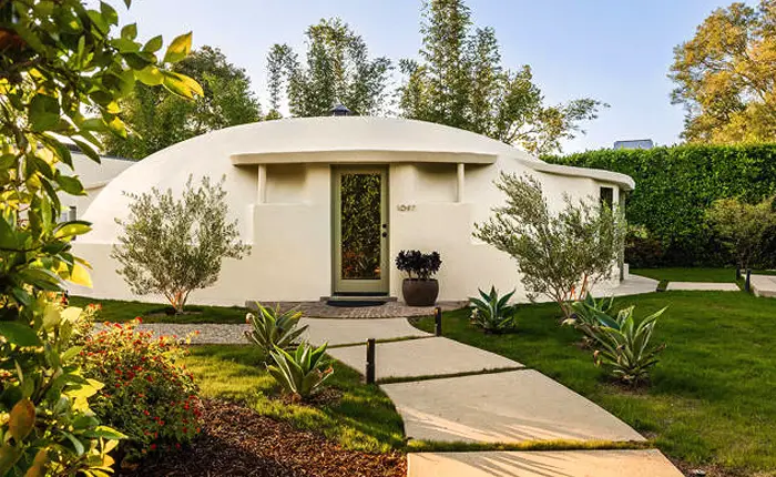 A white, dome-shaped Wallace Neff Shell House with a central door stands amid green grass, plants, and trees, with a winding concrete path leading to the entrance.
