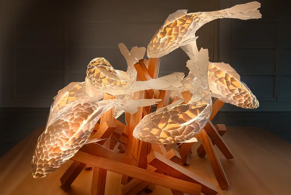 A sculptural fish lamp and sculpture by architect Frank Gehry’s architect fish lamp, featuring textured, translucent koi bodies and colorcore accents, supported by intersecting wooden beams against a neutral indoor background.