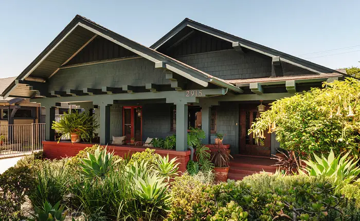 The historic A.C. Tobias Residence is a Craftsman-style house with dark green wood siding, a wide front porch, and exposed beams, surrounded by lush plants and shrubs under a clear sky.
