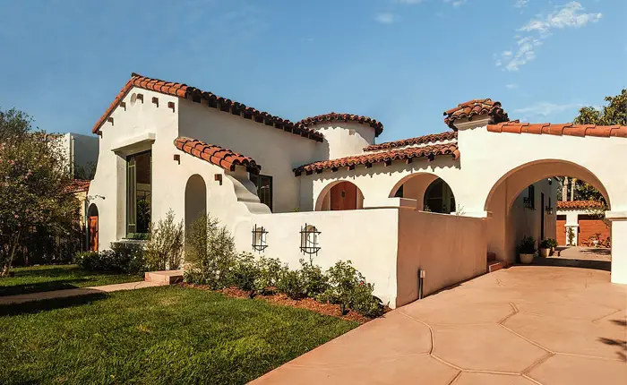 A Historic Spanish Home in Beverly Grove, featuring white stucco walls, red clay tile roofing, arched doorways, and a manicured green lawn under a blue sky.