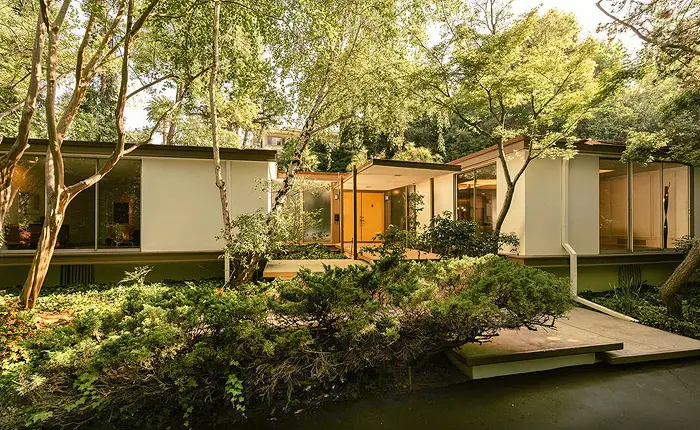 Modern mid-century modern house in Pasadena with large glass windows, designed by David Stringer. Surrounded by lush greenery and tall trees, a pathway leads to the entrance as natural light creates a peaceful, inviting atmosphere.