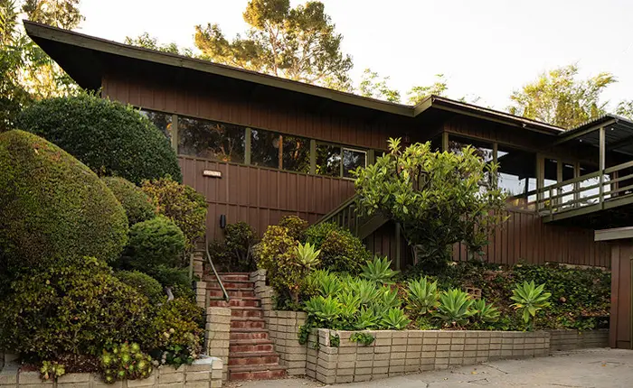 A mid-century modern house in University Hills, with wood siding, large windows, and an angled roof, is surrounded by lush green bushes, succulents, and manicured topiary. A brick staircase leads up to the Robert Pena Rosales Residence entrance.