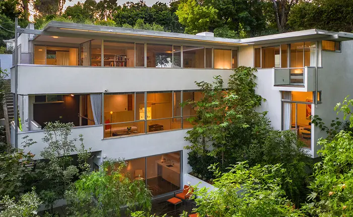 Mid-Century Modern three-story Hendershot House by architect Richard Neutra in Nichols Canyon features large glass windows, surrounded by lush green trees and vegetation. Warm interior lighting contrasts with the cool exterior, creating an inviting atmosphere.