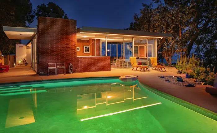 A mid-century modern house, designed by Richard Neutra, features large glass windows and brick walls illuminated at dusk, overlooking a glowing turquoise swimming pool surrounded by patio furniture and trees.