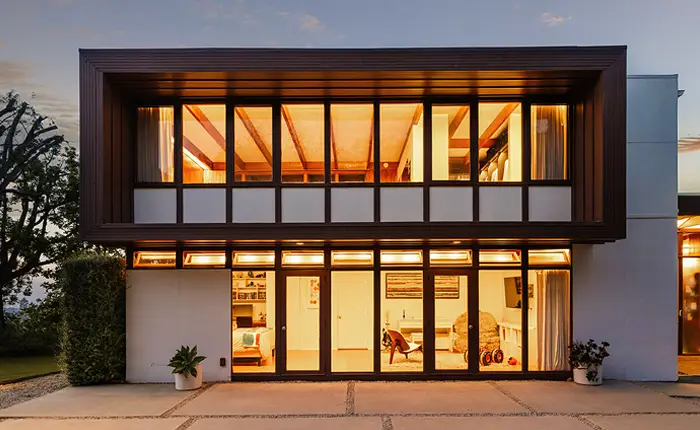Two story Asher House by architect Rodney Walker with large floor-to-ceiling windows illuminated from within at dusk, showcasing a cozy interior. The Mid Century Modern exterior features clean lines, brown framing, and plants near the entrance.