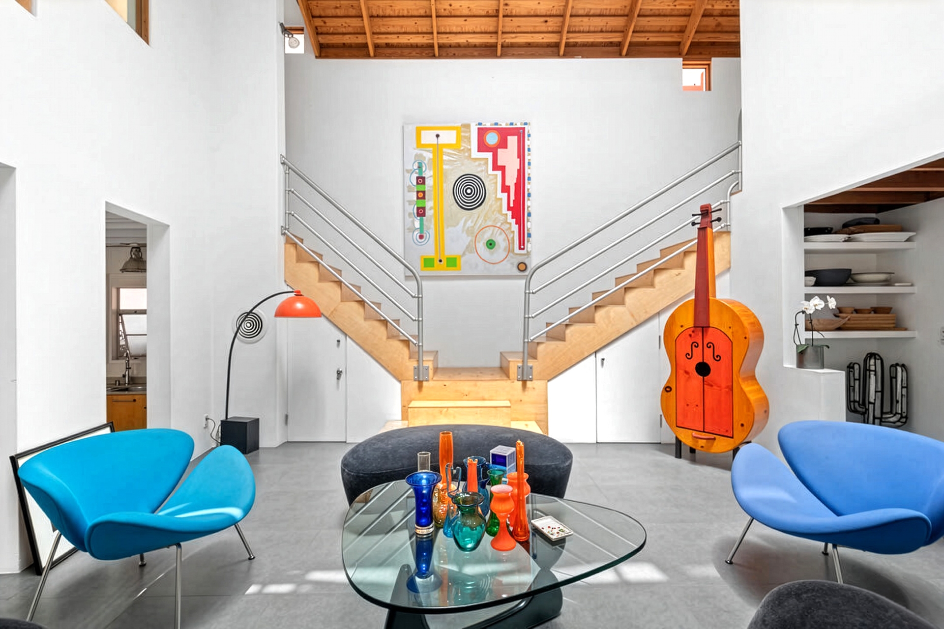 Modern living room at The Caplin House by Frederick Fisher in Venice, featuring blue chairs, a glass coffee table holding colorful vases, and a large, orange upright bass sculpture near a wooden staircase. Abstract artwork hangs on the white wall above the stairs.