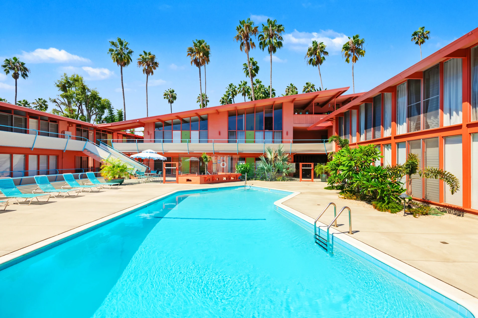 A bright, sunny day at the Hollywood Riviera with an outdoor swimming pool, lounge chairs, palm trees, and coral-colored buildings surrounding the pool area capturing the iconic mid-century modern architecture of Edward Fickett.