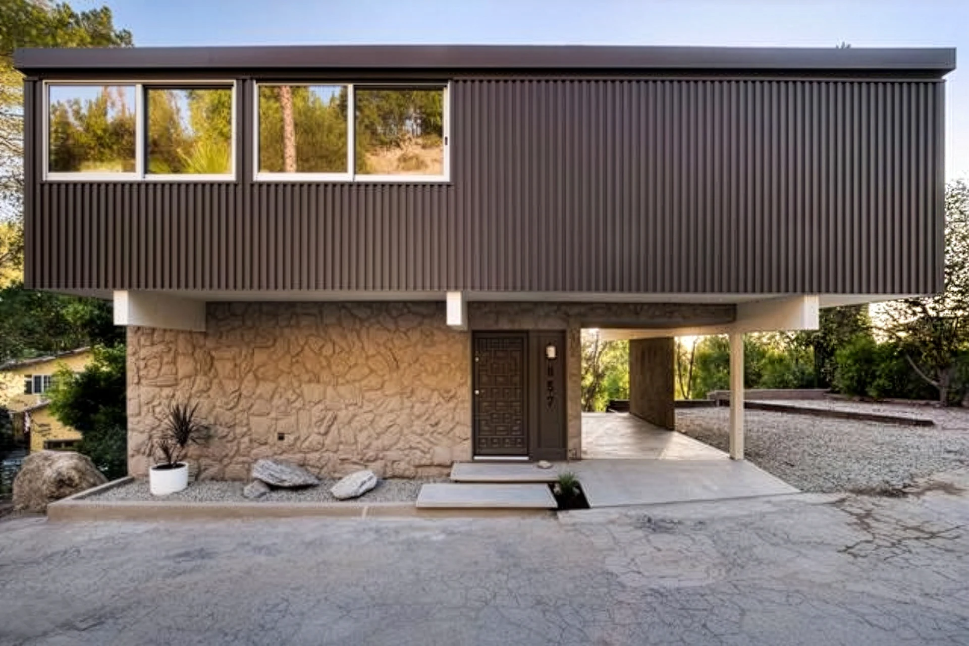 A two-story Mid Century Montecito Heights home with a stone lower level, dark upper level with large windows, a flat roof, and a carport, surrounded by trees and a gravel yard, effortlessly blends Modernist design elements for timeless appeal.